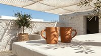 Two terracotta ceramic mugs on white stone surface under woven shade creating shadow patterns on Mediterranean terrace with olive tree and traditional architecture.