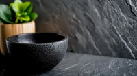 Black textured ceramic bowl with water droplets on dark stone surface, minimalist composition with small green plant in wooden pot, moody atmospheric lighting.