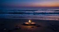 Peaceful beach sunset with glowing candle on sand creating warm light reflection. Ocean waves gently rolling onto shore during golden hour twilight sky.