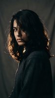 Young woman with dark curly hair in dramatic lighting wearing black coat looking over shoulder with mysterious expression in moody atmospheric setting.