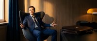 Confident businessman in navy suit sitting in modern leather chair during golden hour lighting in luxury office interior for corporate success concepts.