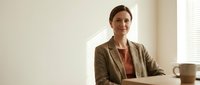 Professional middle-aged Caucasian businesswoman in beige blazer sitting confidently at office desk with natural lighting from window blinds.