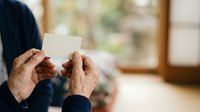 Senior man with wrinkled hands holding blank white card, wearing dark clothing indoors near window. Suitable for business, greeting, invitation, and communication concepts.