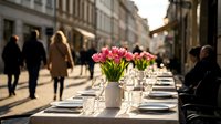 Outdoor dining table with pink flowers in vase, place settings and glasses on urban street with pedestrians walking in background during daytime event or celebration.