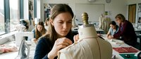 Young female fashion design student working on dress form in bright classroom studio with peers learning garment construction and pattern making skills.
