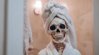 Skeleton in bathroom with white towel wrapped around head, showcasing humorous spa day concept with bone structure visible against neutral beige background.