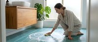 Woman in casual beige shirt cleaning turquoise tile floor with brush in modern bathroom with wooden vanity and potted plant.