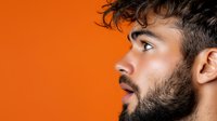 Profile view of young Hispanic man with stylish beard and modern haircut against vibrant orange background, expressing surprise with parted lips.