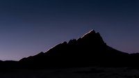 Silhouetted mountain peak at dusk with warm golden light touching the summit against a deep blue twilight sky, suitable for nature and landscape backgrounds.