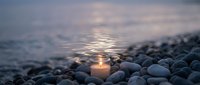 Glowing candle flame surrounded by smooth pebbles on beach shore at twilight with soft ocean waves and peaceful atmospheric lighting for meditation concepts.