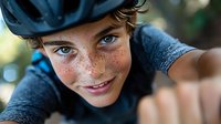 Young Caucasian boy with freckles and bright blue eyes wearing cycling helmet smiles at camera during outdoor activity, close up portrait with natural bokeh background.