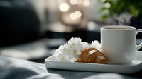 Steaming coffee cup with fresh croissant and white spring flowers on white ceramic plate against blurred background with morning light and cozy atmosphere.