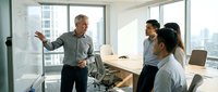 Mature Caucasian male professional presenting business strategy on whiteboard to diverse team in modern high-rise office with city skyline view. Corporate training and workplace collaboration.