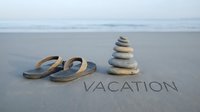 Flip flops and stacked stones on sandy beach with word vacation written in sand, ocean waves in background, summer travel concept.