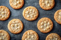 Fresh baked white chocolate macadamia nut cookies arranged in rows on dark textured surface, showing crispy edges and gooey centers with golden brown color.