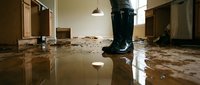 Person in black rubber boots standing in flooded home interior with muddy water covering damaged floor after natural disaster.