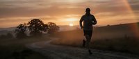 Male athlete jogging on rural road at sunrise, silhouetted against golden sky with misty landscape and mountains in background for fitness and wellness content.