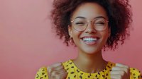 Young African American woman with natural curly hair wearing glasses and yellow polka dot top expressing joy and excitement against pink background.