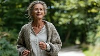 Mature woman with gray hair jogging in park, wearing cozy cardigan over white shirt, enjoying active lifestyle outdoors in natural green setting.