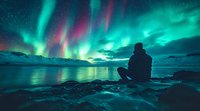 Silhouetted person watching vibrant aurora borealis lights dance across night sky, reflecting in calm water surrounded by snowy mountains and rocky shoreline.