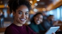 Young African American woman smiling warmly in modern cafe or restaurant setting with friends using tablet in background for social media content.