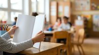 Person with medium skin tone holding open blank notebook in classroom setting with students and teacher in background, suitable for education and learning concepts.