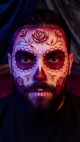 Bearded man with Day of the Dead sugar skull face paint featuring roses and ornate patterns in dramatic purple and red lighting for Halloween celebration.