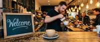 Cozy coffee shop interior with welcome chalkboard sign, latte art cup, and busy barista serving customers in warm ambient lighting.