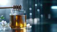 Golden honey dripping from wooden dipper into glass jar, with fresh white cherry blossoms against blurred teal background, natural food photography.
