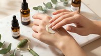 Hands applying natural skincare serum with dropper bottles and eucalyptus leaves on clean white surface for beauty routine and wellness.