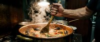 Professional chef cooking traditional paella with seafood in large pan, stirring with wooden spoon, steam rising from hot Spanish rice dish in restaurant kitchen.