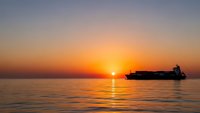 Cargo ship silhouette sailing across calm ocean waters during golden sunset with warm orange and purple sky colors reflecting on sea surface.