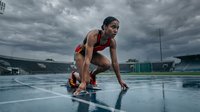 Athletic African American woman sprinter in starting position on wet track under dramatic stormy sky preparing for race at stadium.