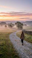 Woman jogging on rural gravel path through misty countryside at sunrise, wearing brown jacket. Scenic landscape with rolling hills, trees, and golden sky for outdoor fitness and wellness.