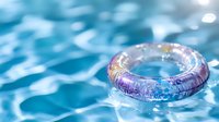 Transparent iridescent swimming pool ring floating on rippling blue water surface with sunlight reflections and bokeh effects creates summer mood.