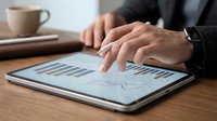 Professional businessman analyzing financial data charts and graphs on digital tablet at wooden desk with coffee cup in modern office workspace.