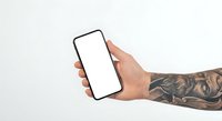Tattooed hand holding blank smartphone with white screen against neutral background for mockup and app presentation purposes.