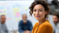 Young Caucasian businesswoman in yellow sweater smiling confidently during office meeting with colleagues, focus on female professional with natural makeup and curly hair.