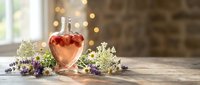 Heart-shaped glass bottle with pink liquid surrounded by lavender flowers and white blooms on wooden table near window with warm bokeh lights.