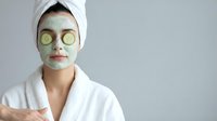 Young woman in white bathrobe with green facial mask and cucumber slices on eyes for spa skincare treatment and relaxation at home wellness routine.