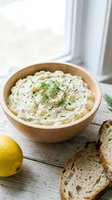Creamy potato salad with fresh dill and black pepper in wooden bowl, served with toasted bread and lemon on white wooden table near window.