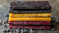 Stack of dark, milk and ruby chocolate bars with chocolate chips scattered on rustic surface, macro food photography showing texture and layers.