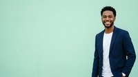 Young man with dark skin tone and black curly hair wearing navy blazer and white shirt, smiling confidently against mint green background. Corporate portrait for business, professional headshot, and marketing materials.