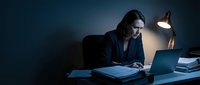 Focused businesswoman working late at night on laptop computer in dark office with desk lamp illumination for overtime productivity and dedication.