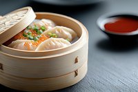 Asian dumplings with salmon roe in traditional bamboo steamer, garnished with green onions, served with red sauce on dark stone background.