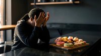 Young African American woman feeling overwhelmed by unhealthy food choices, struggling with diet decisions in modern kitchen setting.