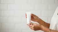 Woman with medium skin tone holding white cosmetic bottle against white brick wall background for skincare product marketing.