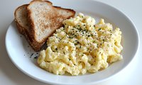 Creamy scrambled eggs seasoned with black pepper served on white plate with toasted bread slice, close up view of homestyle breakfast preparation.