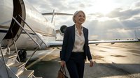 Mature woman in business attire walking on airport tarmac toward private jet, carrying leather briefcase, suitable for corporate travel and executive lifestyle concepts.