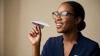 Professional African American businesswoman in glasses holding paper airplane, smiling with optimism and creativity in modern office setting.